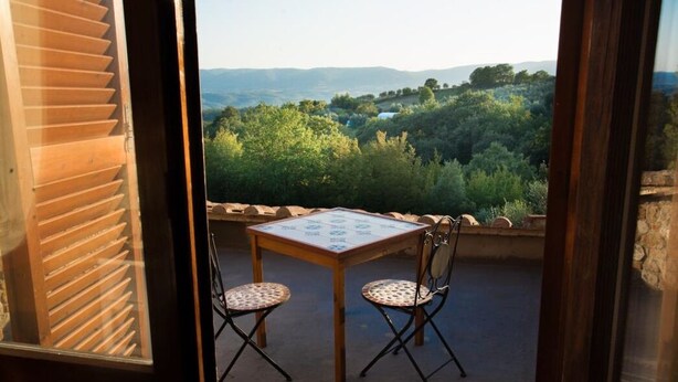 A balcony with a table and two chairs, overlooking a lush landscape with trees and hills.