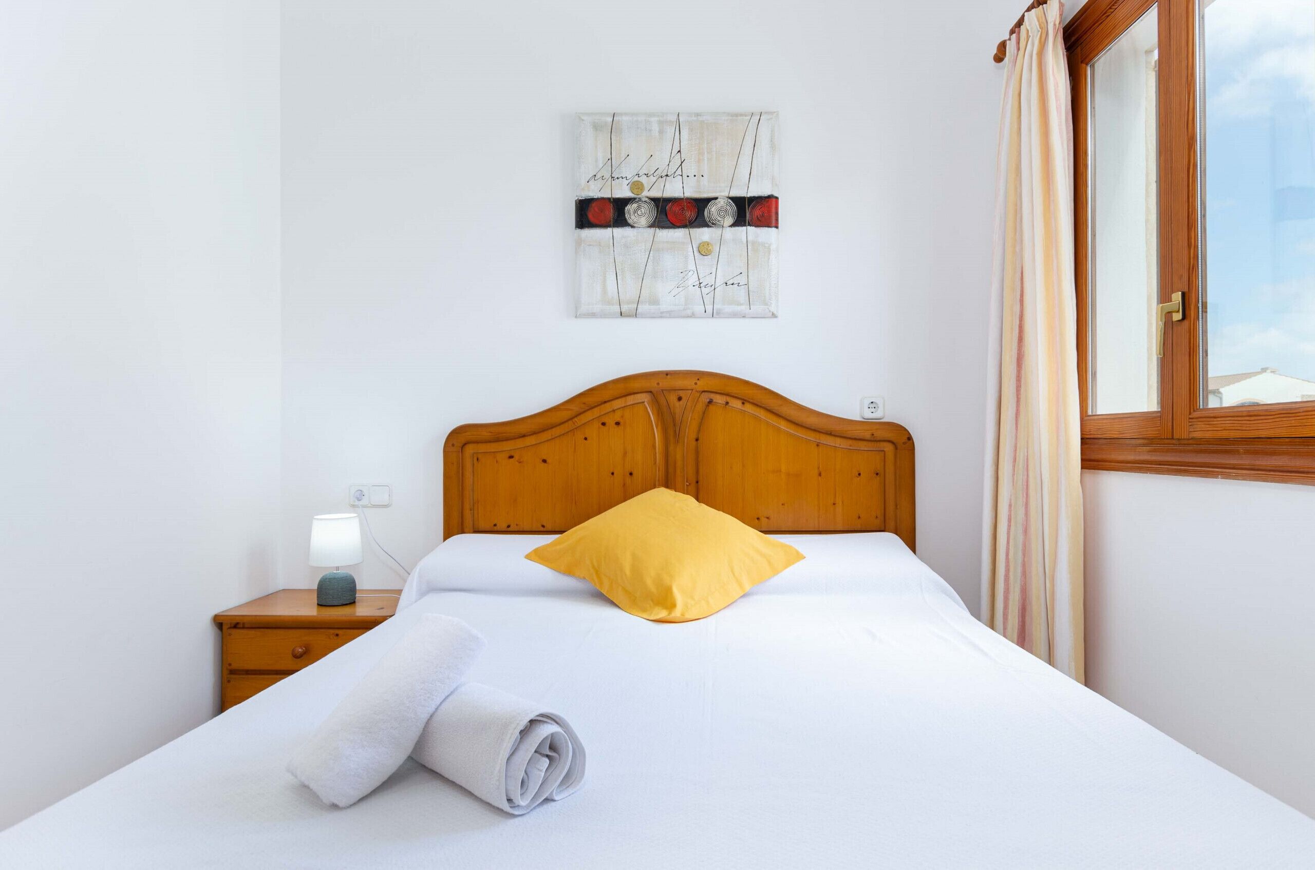 Una cama bien hecha con cabecera de madera, un almohadón amarillo y una toalla blanca enrollada en la mesita de noche.