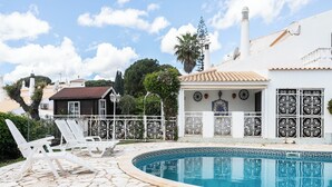 A pool area with white chairs, a tiled deck, and a white building with decorative window grills. - Feta Villa, Pool, BBQ (Olhos de Água)