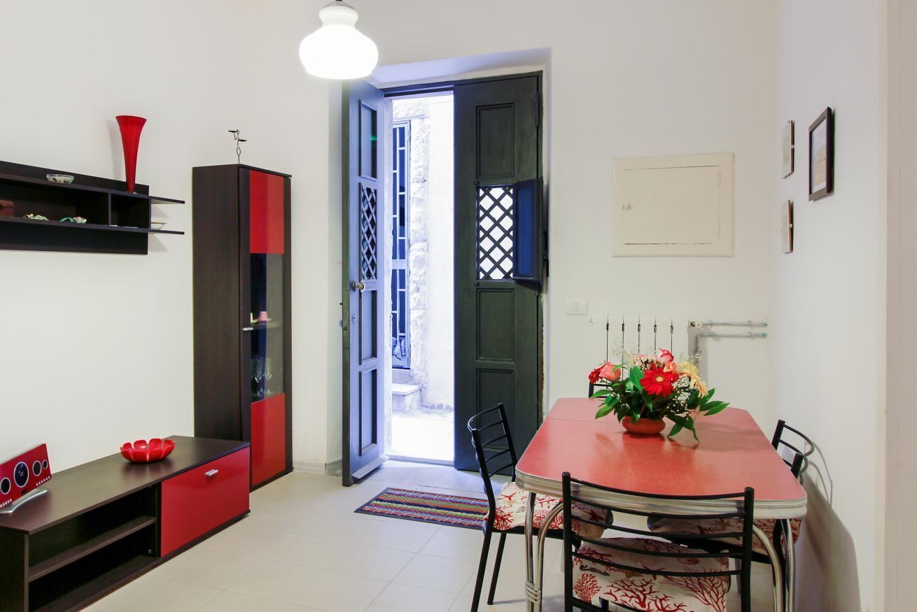 Ein Essbereich mit einem roten Tisch, Stühlen und einer Blumenvase. Es gibt einen schwarzen Schrank mit roter Akzentfarbe, eine rote Vase auf einem Regal und eine Tür, die nach draußen führt.