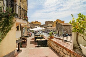 Une rue étroite avec des terrasses, des bâtiments en pierre et, au fond, une structure qui ressemble à un château.