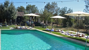 Une piscine entourée d’arbres et d’un bâtiment à toit de tuiles, avec des chaises longues et des parasols à proximité.