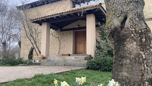 Un bâtiment en pierre doté d’une porte en bois et de colonnes, entouré de verdure et de jonquilles.