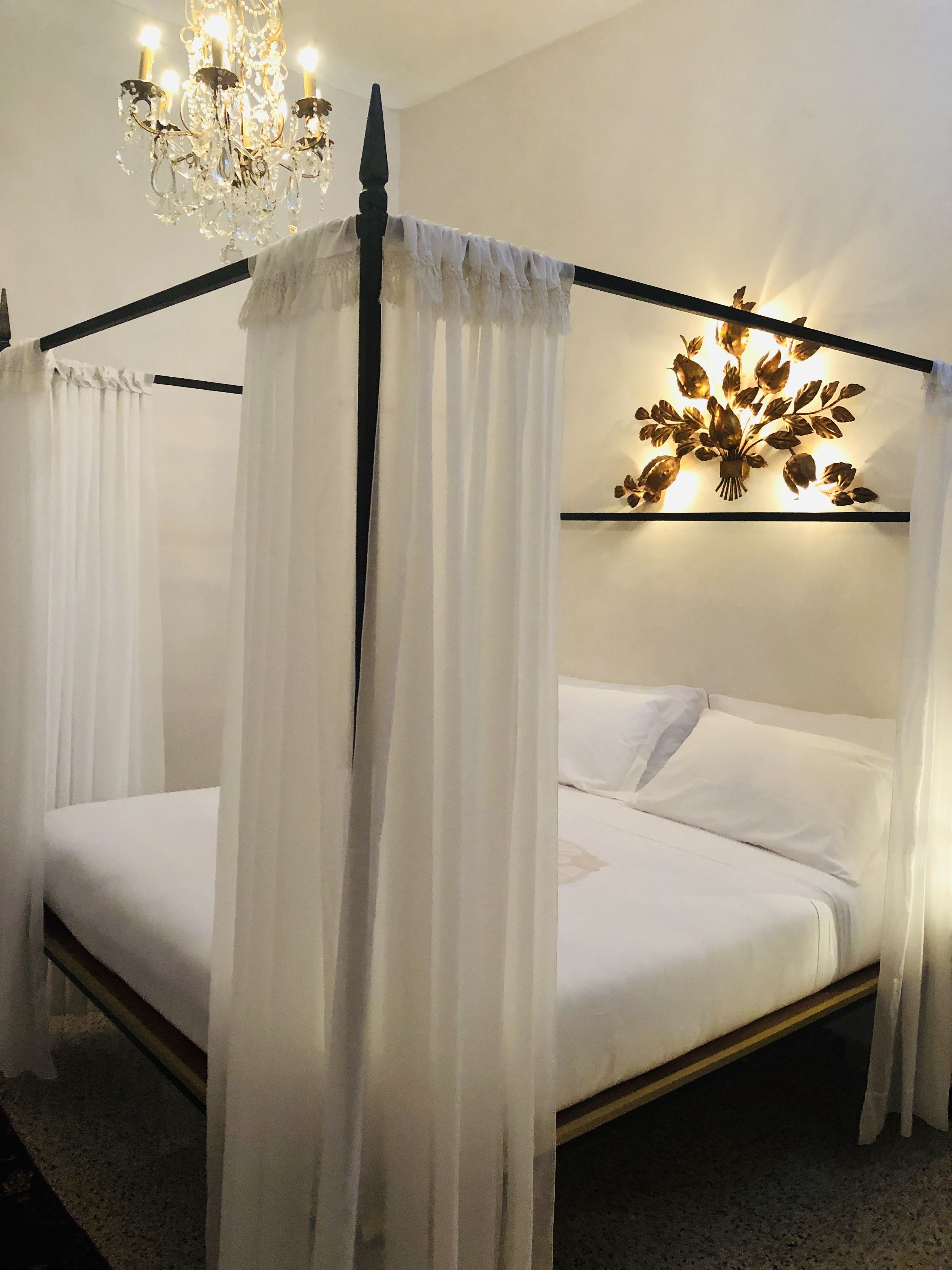 A four-poster canopy bed with white linens and sheer curtains, a chandelier, and a wall-mounted light fixture with a floral design.