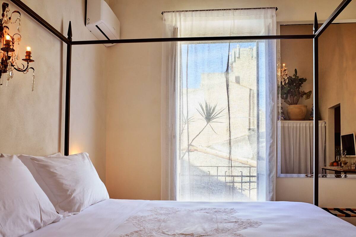 Ein Schlafzimmer mit einem Himmelbett, einem großen Fenster mit Blick auf Gebäude und einem Blumentopf auf dem Fensterbrett.