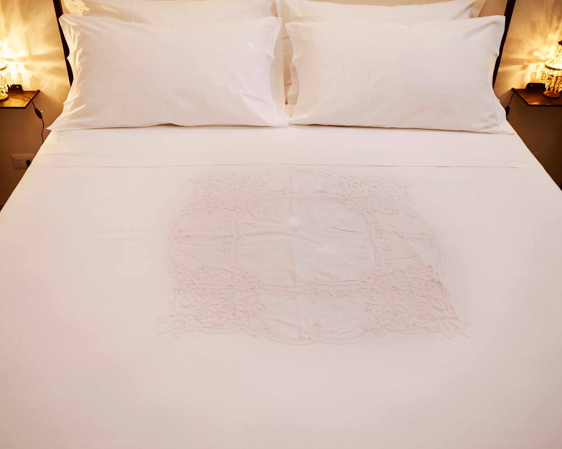 A neatly made bed with a white bedspread and pillows, a decorative embroidered design on the bedspread, and two bedside lamps on either side.