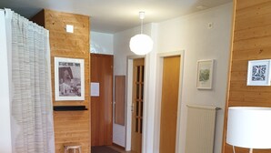 Een kamer met een houten vloer, een tafel met stoelen, een kruk, een lamp op een statief en een deur die leidt naar een andere kamer.