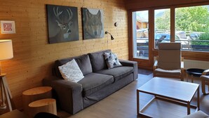 Een gezellige kamer met een houten wand, een grijze bank, een klein tafeltje en een raam met uitzicht op auto’s en groen.
