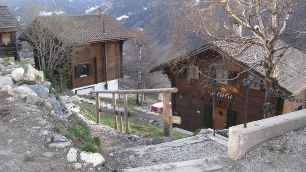 A rustic mountain cabin with a wooden exterior, a stone pathway leading to the entrance, and a view of snow-capped mountains in the background. - Outdoor parking space - close to the cottage (at the start of the stairs) (St-Luc)