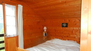 A wooden cabin room with a bed, a nightstand, a lamp, and a window with curtains. - Outdoor parking space - close to the cottage (at the start of the stairs) (St-Luc)