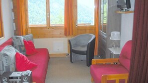 A cozy room with a red sofa, a grey armchair, a small table with a radio, and a TV on a stand. - Outside : Living on the 3rd floor of an apartment house, in Chandolin, for 2-4 persons, balcony with (Chandolin)