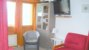A room with a wooden cabinet, a television, a red chair, a grey chair, and a window with curtains. - Outside : Living on the 3rd floor of an apartment house, in Chandolin, for 2-4 persons, balcony with (Chandolin)