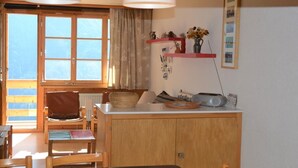 Een knus ingerichte kamer met een houten kast, een eettafel met stoelen, een raam met gordijnen en een plank met decoratieve spullen.