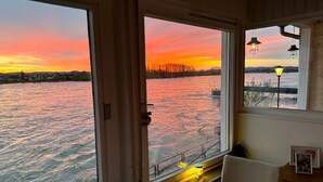 Una habitación con un ventanal grande que da a un cuerpo de agua durante el atardecer, un escritorio pequeño con una silla y una lámpara.