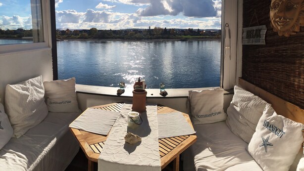 Una habitación acogedora con vista a un lago, una mesa de madera y un sofá con almohadas 'Sommerbrise'.