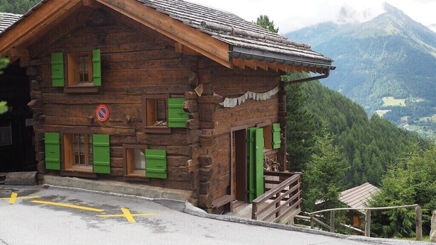 Ein rustikales Holzhaus mit grünen Fensterläden, einem Balkon und Blick auf eine gebirgige Landschaft.