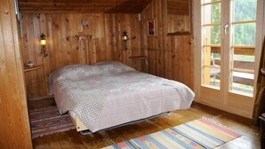 Ein Schlafzimmer einer Holzhütte mit einem Bett, einem Nachttisch, einer Wandlampe und einem Fenster mit Blick ins Freie.