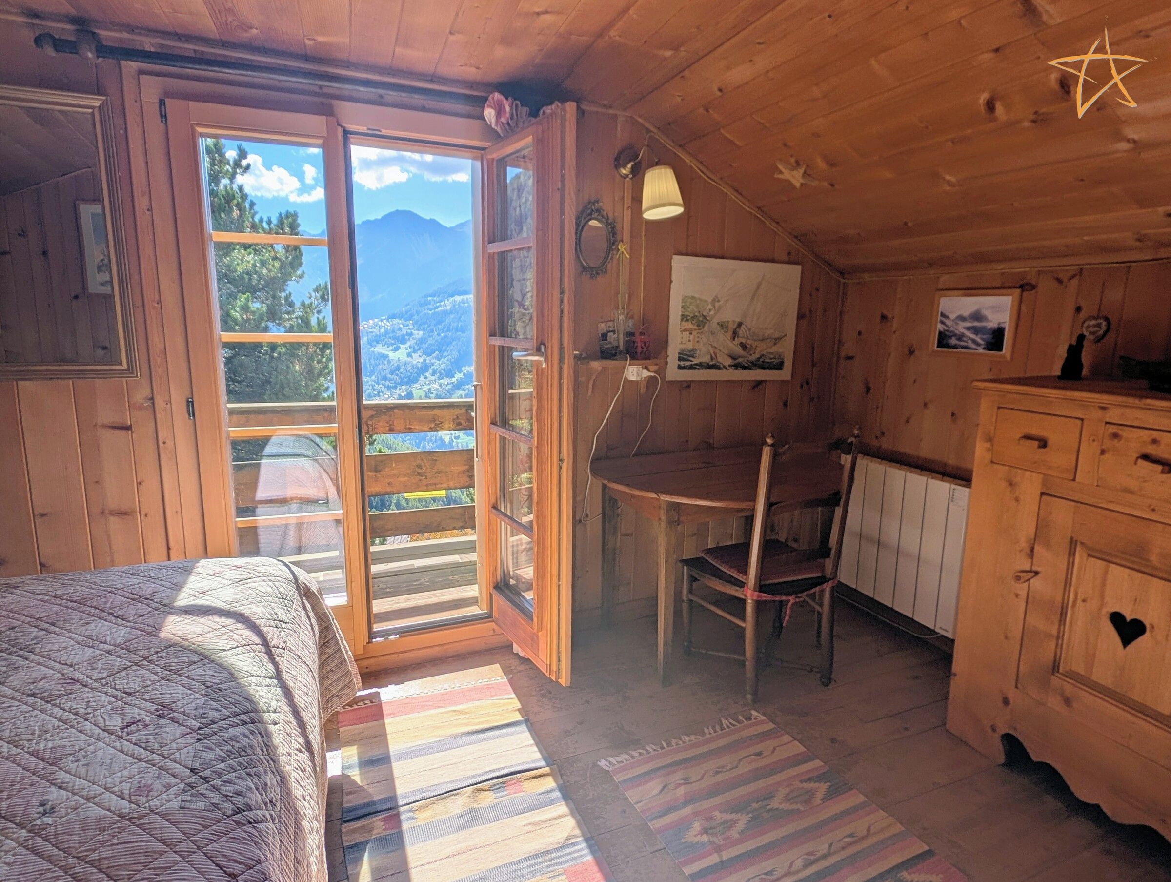 Een houten blokhut van binnen met een bed, een bureau, een stoel en een uitzicht op bergen door een groot raam.