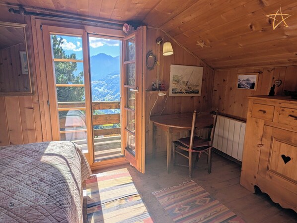 A wooden cabin interior with a bed, a desk, a chair, and a view of mountains through a large window. - Outside: individual chalet on 3 floors for max. 5 people in the center of the old village with direc (St-Luc)