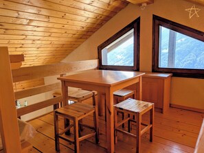 A wooden interior with a sloped ceiling, a table, two barstools, and two windows offering a view of a mountainous landscape.