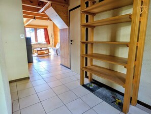 Un pasillo con una estantería de madera, suelo de baldosas y una puerta que da a una zona de estar con un sofá y una mesa.