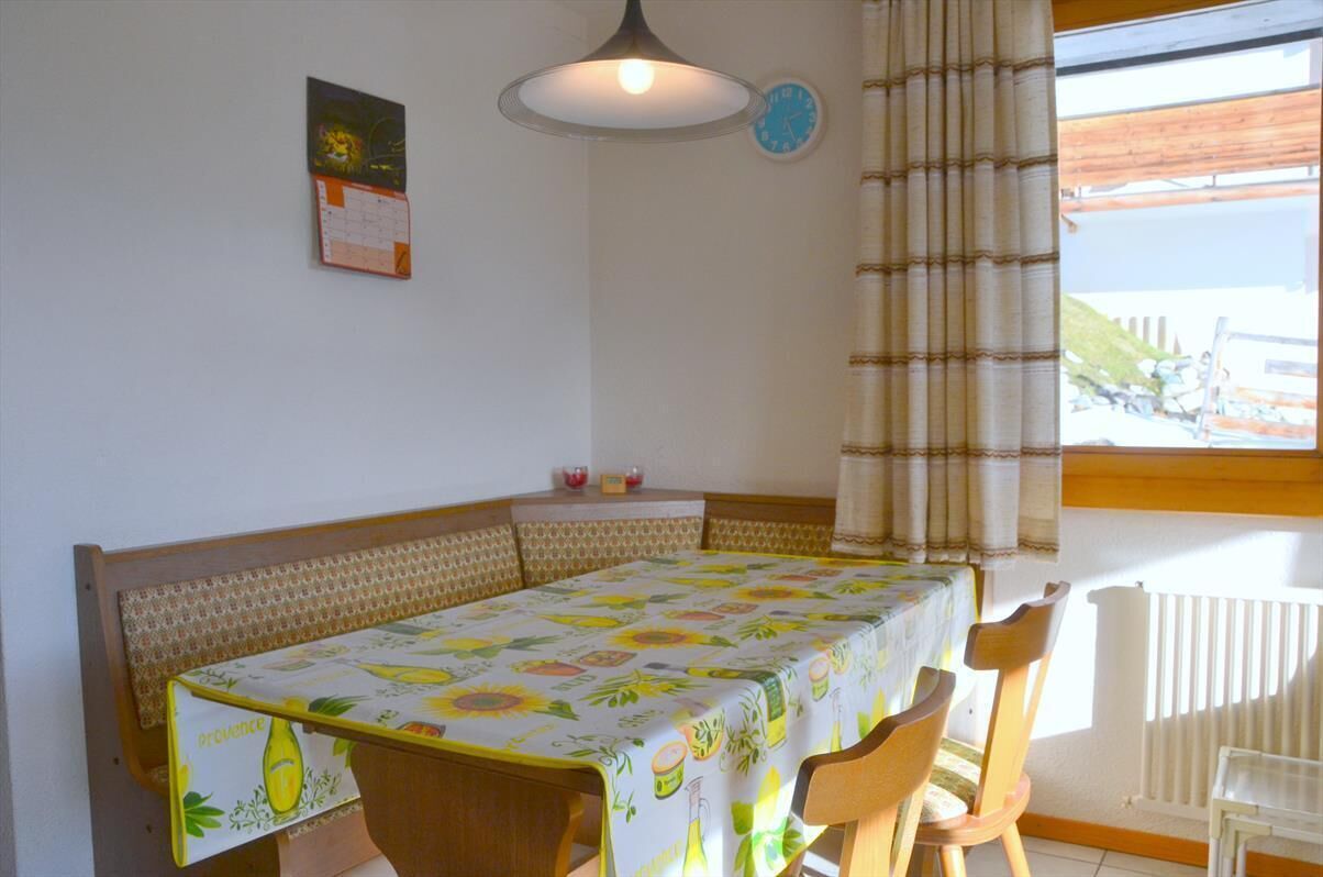 A dining area with a table covered in a fruit-patterned tablecloth, wooden chairs, a bench, a hanging light fixture, a wall clock, and a window with curtains.