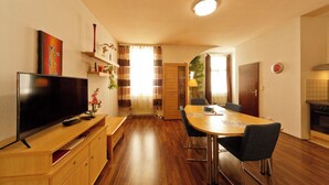 A modern living room with a flat-screen TV, wooden furniture, a dining table with chairs, and a kitchen area with a stove. - Apartment No. 03 in Vienna near Schönbrunn/Westbahnhof | Keyless Check-In (Rudolfsheim-Fünfhaus)