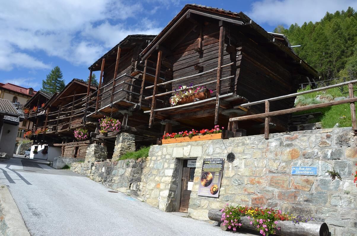 A rustic wooden building with stone walls, flower boxes, and a sign indicating 'Rue des Lavandieres'.