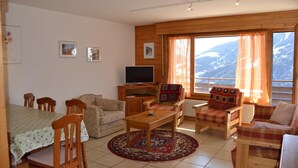 A cozy living room with a TV, wooden furniture, a dining table, and large windows with curtains.