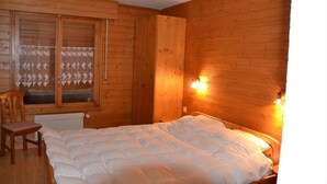 A wooden cabin room with a bed, a chair, and a window with lace curtains.