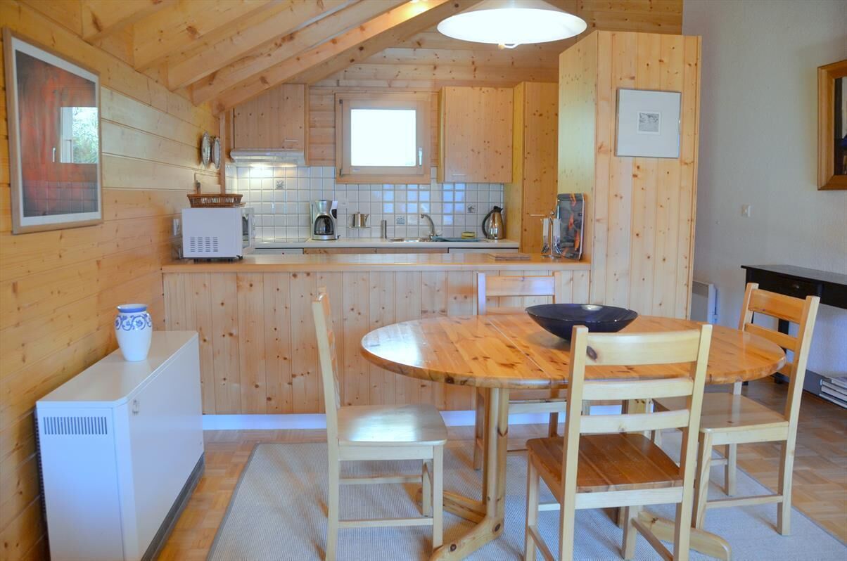 Un interior de madera con una zona de cocina que incluye una mesa redonda de madera, cuatro sillas, un refrigerador blanco y un microondas.