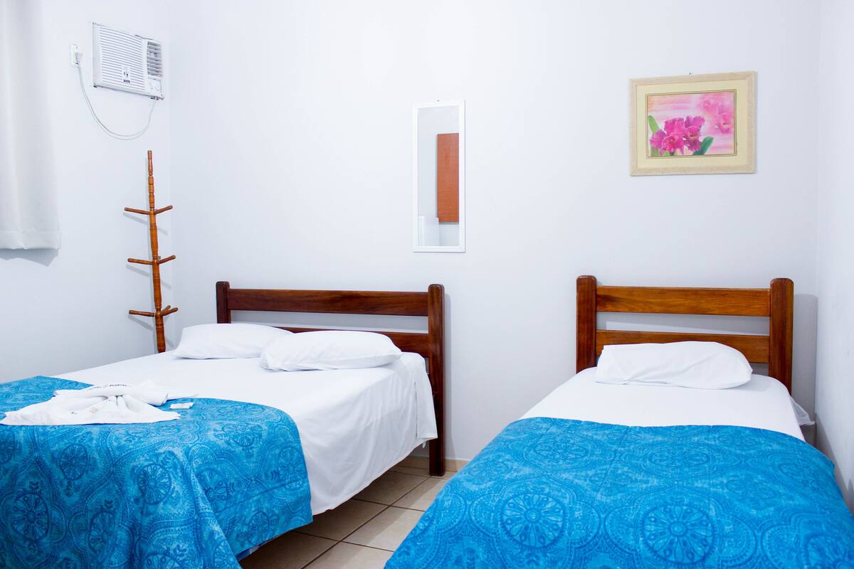 A hotel room with two single beds, a wooden headboard, a wall-mounted air conditioner, a mirror, and a framed picture of flowers.