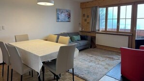 A dining area with a table and chairs, a sofa, and a window with curtains. - Outside: 3-room apartment for 5 people on the lower floor, balcony -Interior: 90 m2, closed entrance (St-Luc)