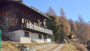 A rustic chalet with a balcony, stone foundation, and a dirt path leading up to it.