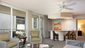 Ein Hotelzimmer mit Sitzecke, einem Glastisch, einer Bar mit zwei Barhocker und einem Deckenventilator.
