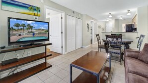 A living room with a flat-screen TV, a coffee table, a sofa, and a dining area with a glass-top table and chairs. - Surf Beach 203 - Gulf Front Condo - ADA (Treasure Island)