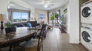 A spacious living area with a dining table, chairs, a sofa, and a balcony with a view. - Surf Beach 203 - Gulf Front Condo - ADA (Treasure Island)
