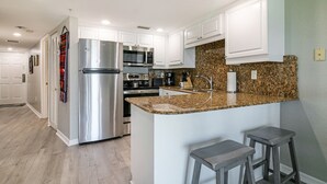 Una cocina moderna con armarios blancos, electrodomésticos de acero inoxidable, encimeras de granito y dos taburetes de bar.
