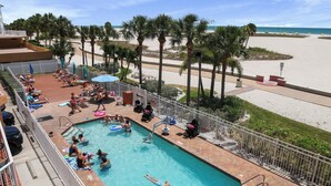 A pool area with a view of the beach, palm trees, and a fenced deck with lounge chairs and umbrellas. - Surf Beach 305 - Spectacular Sunsets - Beach Front (Treasure Island)