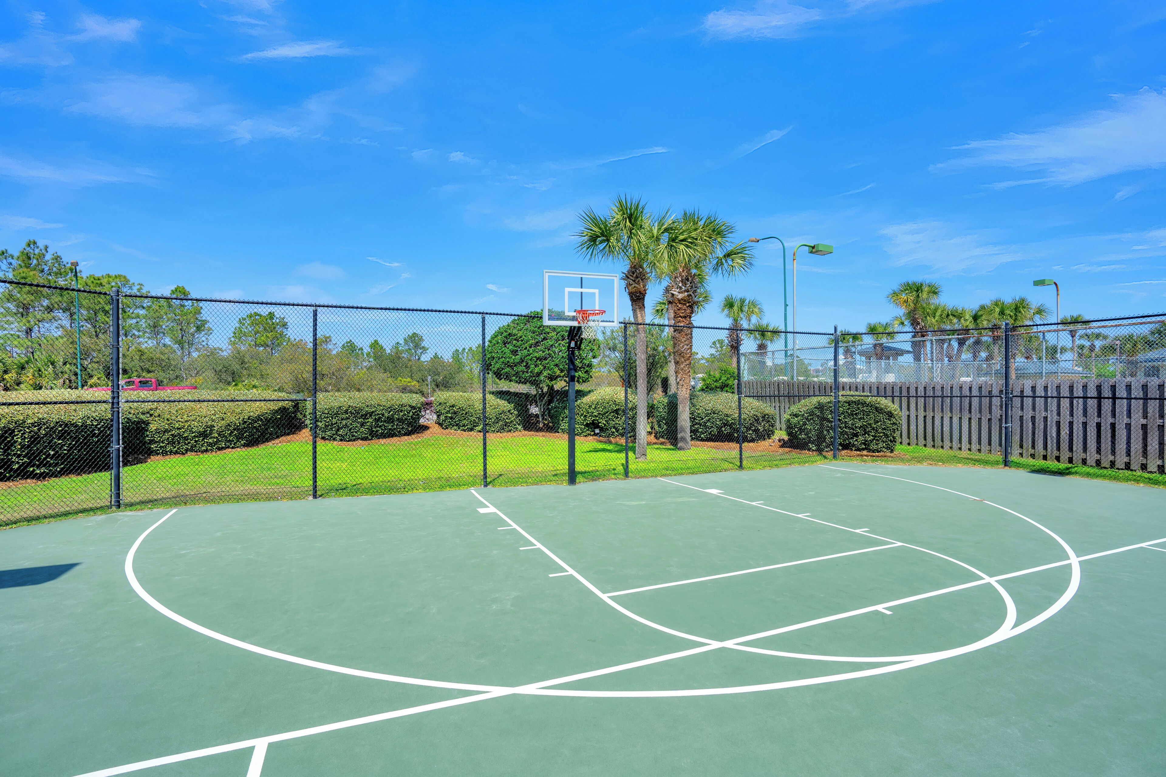 Un campo da basket all'aperto con superficie verde, circondato da una recinzione a rete metallica, palme e un cielo azzurro intenso.