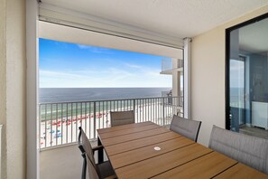 A balcony with a wooden table and chairs, overlooking a beach with umbrellas and a clear sky.