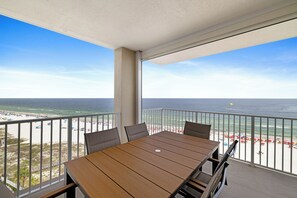 A balcony with a wooden table and chairs, overlooking a beach with umbrellas and a clear view of the ocean.