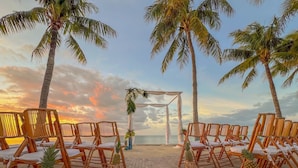 Une cérémonie de mariage organisée sur la plage, avec des chaises en bois et une arche blanche, des palmiers et une vue sur le coucher de soleil.
