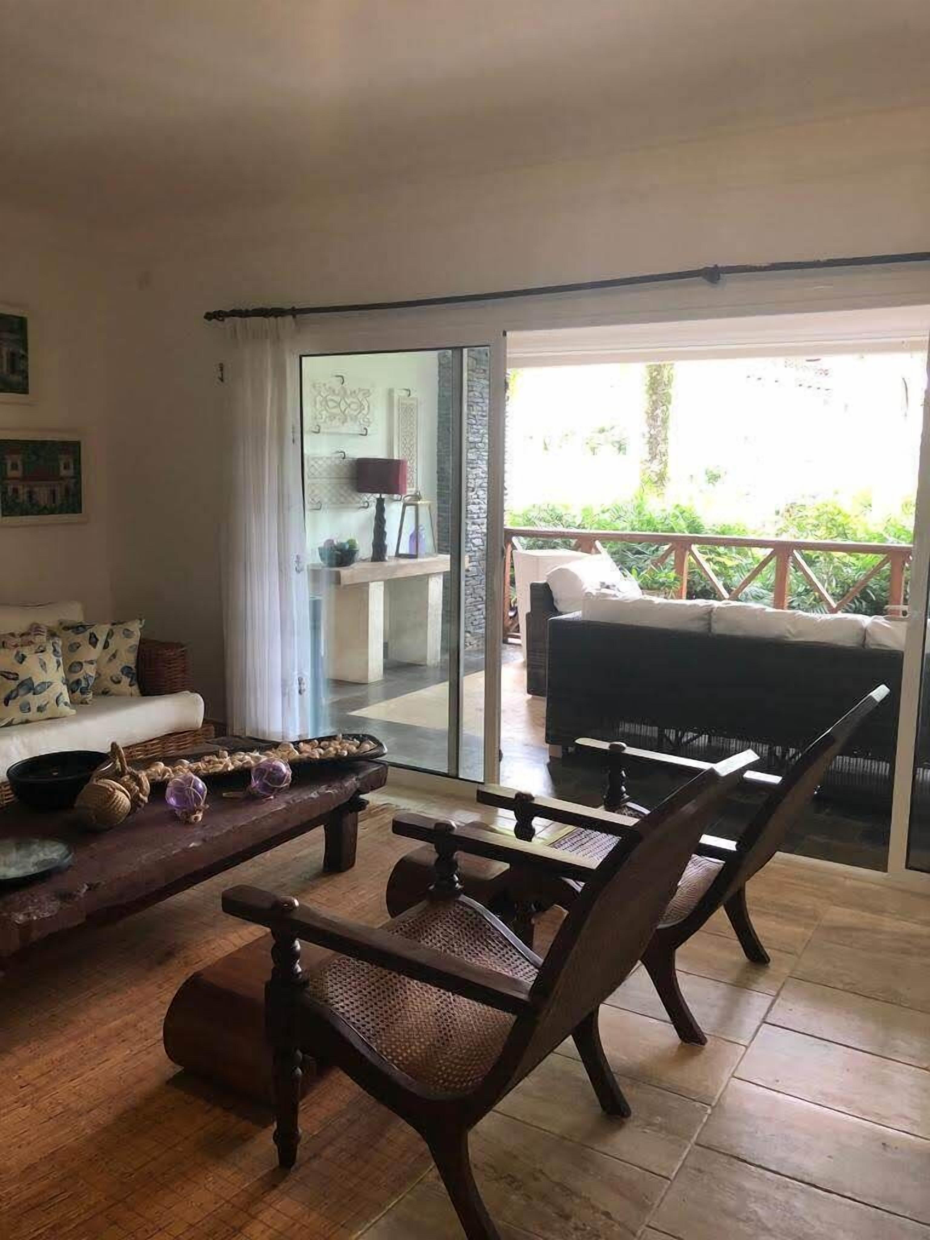 Sala de estar com sofá, mesa de centro de madeira e duas cadeiras. Há uma porta de vidro que dá para uma varanda com vista para áreas verdes.