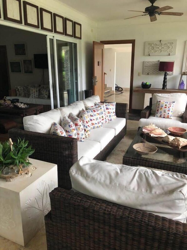 A living room with a white sofa, wicker armchairs, and a coffee table. There are framed pictures on the wall, a ceiling fan, and a door leading to another room.