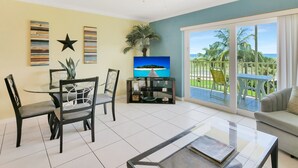 A living room with a dining area, a TV, and a balcony with a view of the ocean.