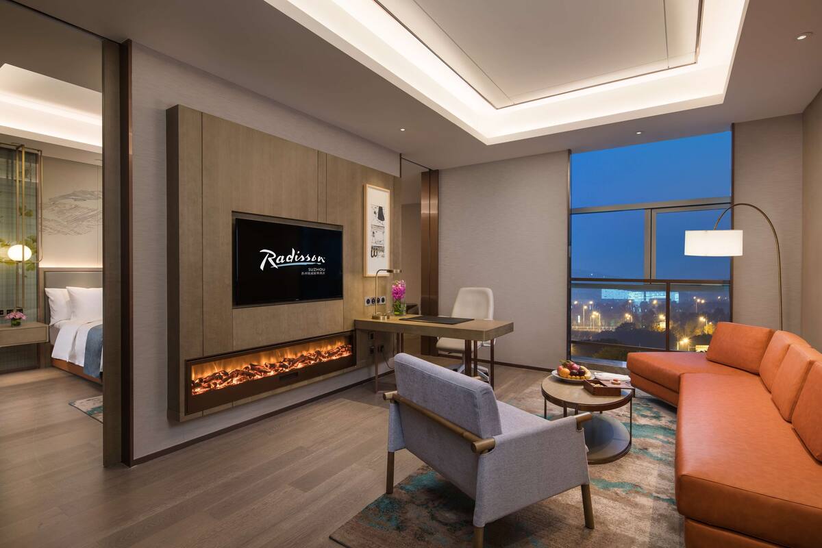 A modern hotel room with a flat-screen TV, a fireplace, a sofa, a dining area, and a large window with a city view.