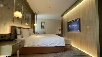 A modern hotel room with a large bed, bedside tables, a flat-screen TV mounted on the wall, and ambient lighting.