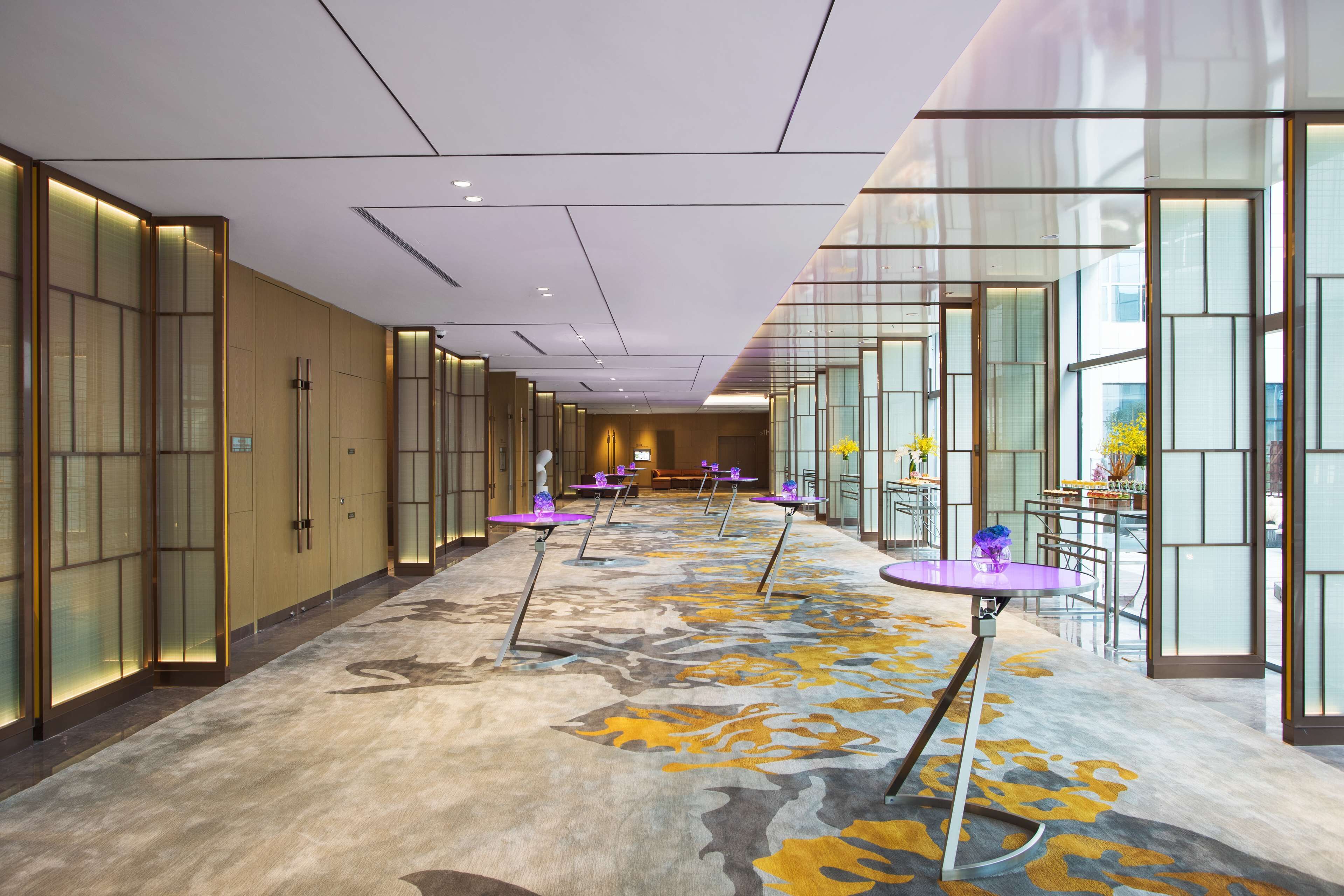 A hotel corridor with a patterned carpet, glass partitions, and purple tables with chairs.