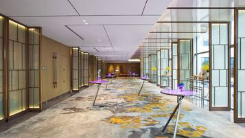 A hotel corridor with a patterned carpet, glass partitions, and purple tables with chairs.